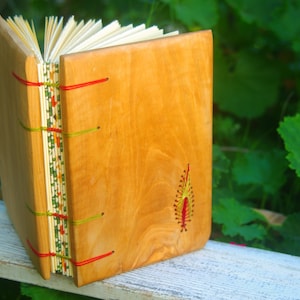 May include: A wooden journal with a natural wood cover and red and green stitching. The journal has a small embroidered design on the front cover.