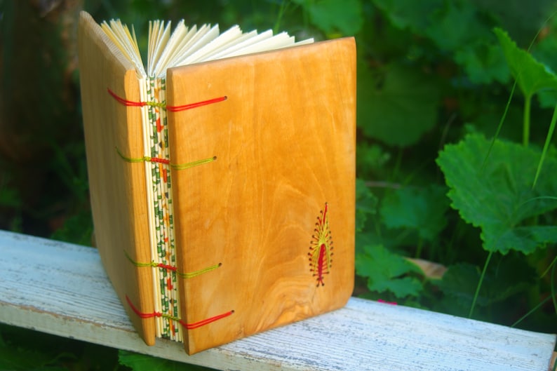 May include: A wooden journal with a natural wood cover and red and green stitching. The journal has a small embroidered design on the front cover.