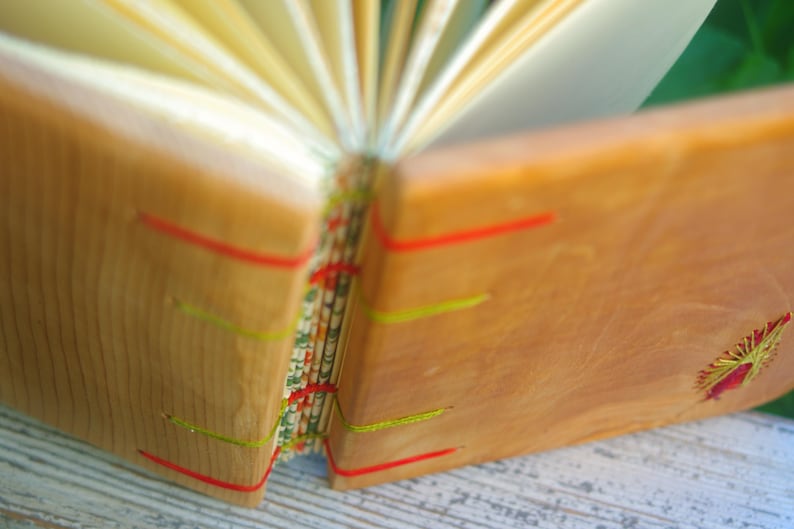 May include: A wooden journal with a natural wood cover and colorful string accents. The journal is open to reveal the pages inside.