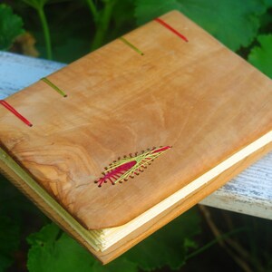 May include: A wooden journal with a natural wood cover and red and green stitching. The cover has a decorative design stitched into it.