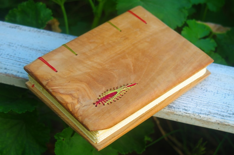 May include: A wooden journal with a natural wood cover and red and green stitching. The cover has a decorative design stitched into it.