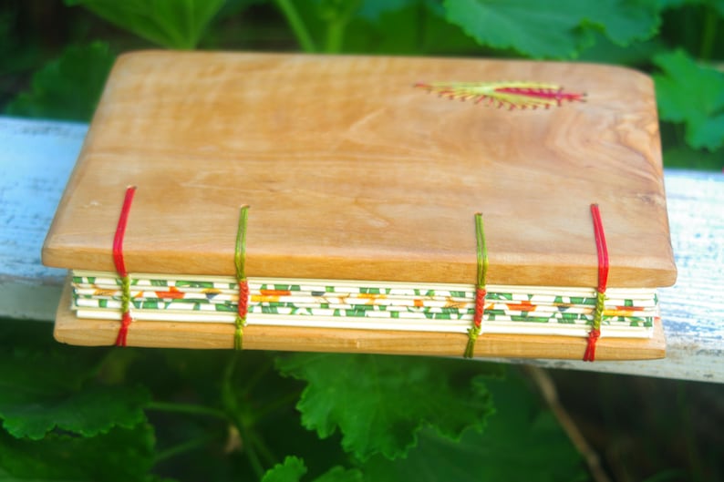 May include: A wooden journal with a natural wood cover and a decorative stitched design. The journal has a green, red, and white patterned paper spine.
