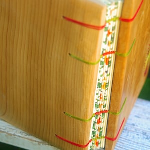 May include: A wooden book with a decorative spine featuring a colorful pattern of red, green, and white. The book is bound with red and green thread.