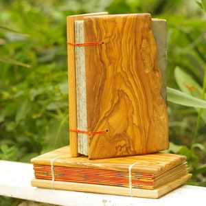 May include: Two handmade journals with wooden covers. The top journal has a natural wood cover with a red string closure. The bottom journal has a red, orange, and yellow patterned cover with a natural wood cover.
