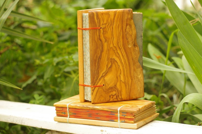 May include: Two handmade journals with wooden covers. The top journal has a natural wood cover with a red string closure. The bottom journal has a red, orange, and yellow patterned cover with a natural wood cover.