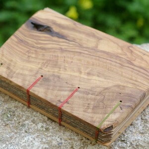 Op de afbeelding: Een handgemaakt dagboek met een houten omslag van olijfhout. De omslag heeft een natuurlijke houtnerf en een knoop in het hout. Het dagboek is gebonden met rood en groen touw.