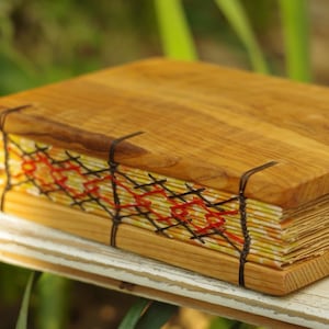 Peut inclure: Un journal artisanal avec une couverture en bois et une reliure colorée et à motifs. La reliure est faite de fil noir et de points rouges.