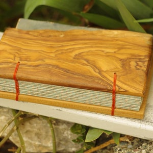 May include: A handmade journal with a wooden cover made from olive wood. The cover has a natural wood grain pattern and is secured with red string. The journal has a light blue and white patterned paper cover.