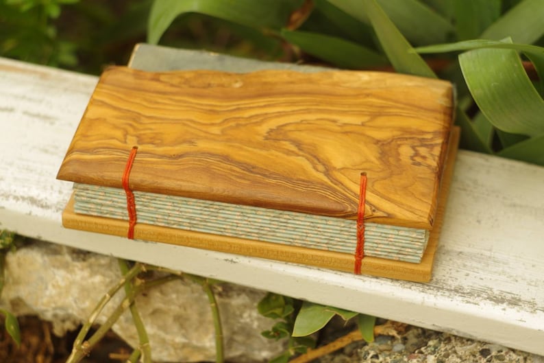 May include: A handmade journal with a wooden cover made from olive wood. The cover has a natural wood grain pattern and is secured with red string. The journal has a light blue and white patterned paper cover.