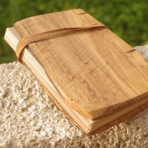 May include: A wooden journal with a natural wood cover and a leather strap closure. The journal is closed and lying on a light-colored surface.