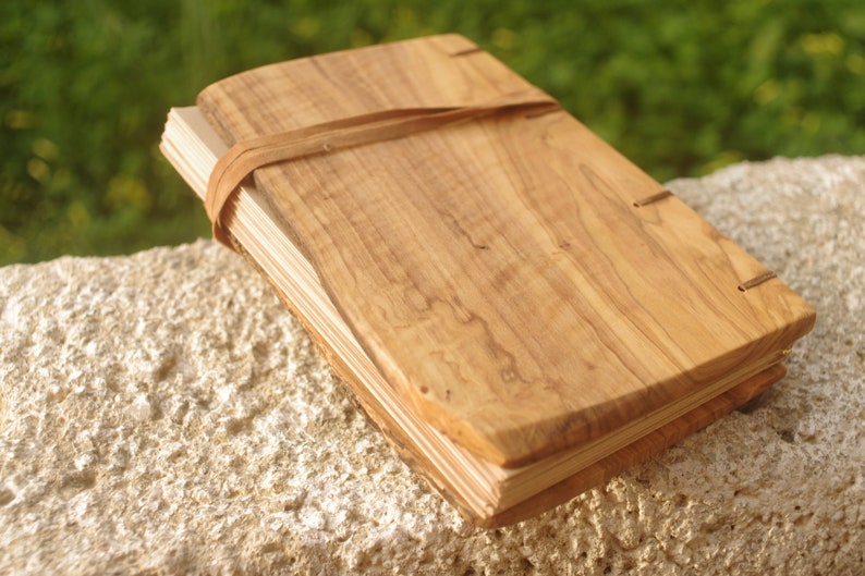 May include: A wooden journal with a natural wood cover and a leather strap closure. The journal is closed and lying on a light-colored surface.