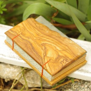 May include: A small, handmade journal with a wooden cover made from olive wood. The cover has a natural wood grain pattern and is secured with red string.