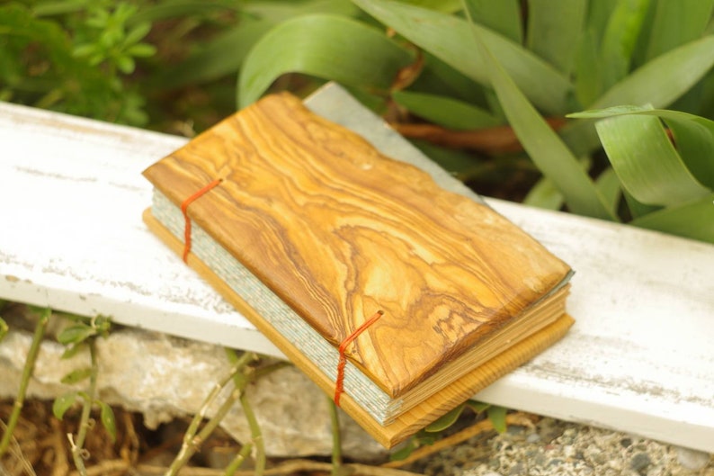 May include: A small, handmade journal with a wooden cover made from olive wood. The cover has a natural wood grain pattern and is secured with red string.