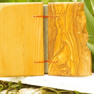 May include: A handmade journal with a wooden cover. The front cover is made of light wood with a smooth finish. The back cover is made of olive wood with a natural, rustic finish. The journal is bound with red string.