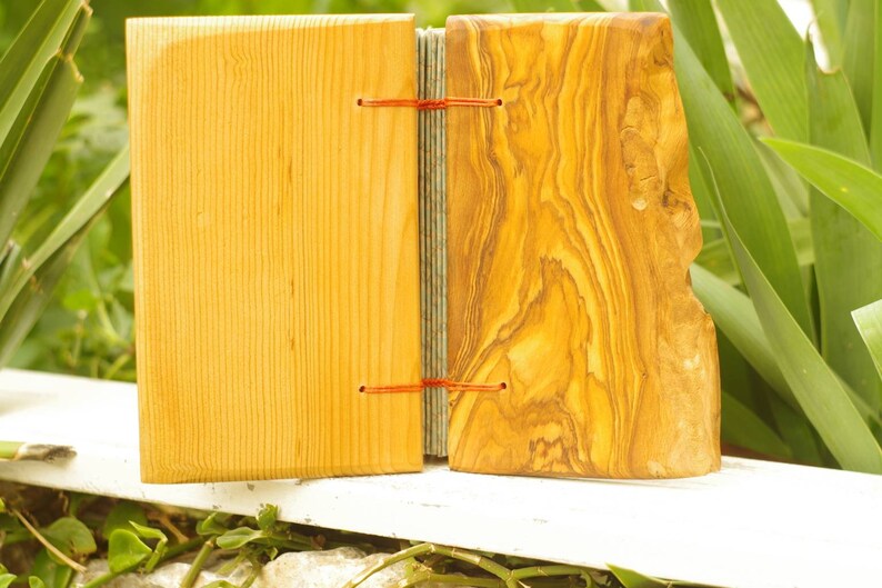 May include: A handmade journal with a wooden cover. The front cover is made of light wood with a smooth finish. The back cover is made of olive wood with a natural, rustic finish. The journal is bound with red string.