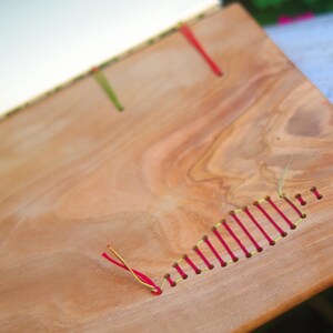 May include: A wooden book cover with red and yellow thread sewn into the surface. The wood has a natural grain pattern.