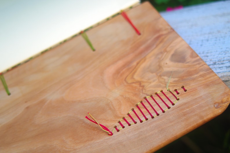 May include: A wooden book cover with red and yellow thread sewn into the surface. The wood has a natural grain pattern.