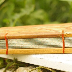May include: A handmade journal with a wooden cover and red string binding. The cover is made of light brown wood with a natural grain pattern. The pages are bound together with a red string and are visible through the open cover.
