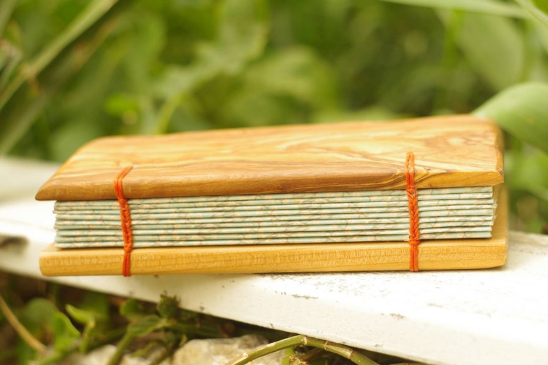 May include: A handmade journal with a wooden cover and red string binding. The cover is made of light brown wood with a natural grain pattern. The pages are bound together with a red string and are visible through the open cover.