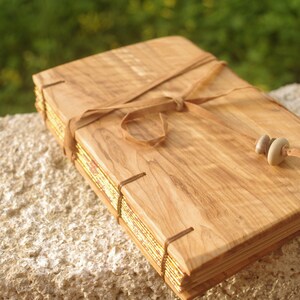 May include: A wooden journal with a leather strap closure and gold leaf accents. The journal is tied with a brown leather strap and has two small stones as a bookmark.