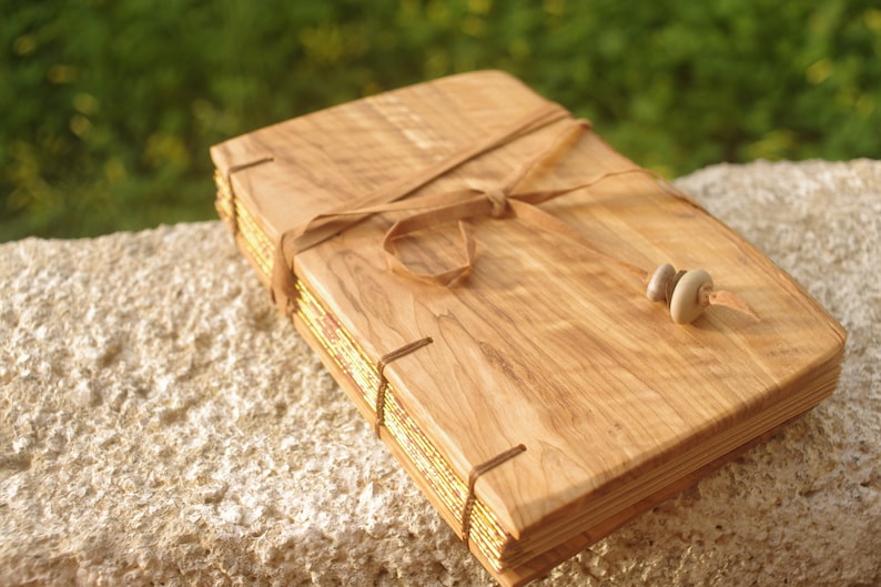 May include: A wooden journal with a leather strap closure and gold leaf accents. The journal is tied with a brown leather strap and has two small stones as a bookmark.