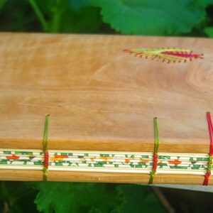 May include: A wooden journal with a natural wood cover and a decorative floral design embroidered on the front. The journal is bound with red, green, and white thread.