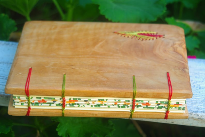 May include: A wooden journal with a natural wood cover and a decorative floral design embroidered on the front. The journal is bound with red, green, and white thread.