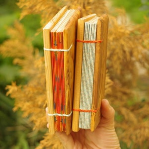 May include: Two handmade journals with wooden covers. The journal on the left has a red and orange patterned paper cover. The journal on the right has a white and gray patterned paper cover. Both journals are bound with red string.