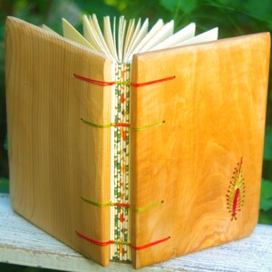 May include: A wooden journal with a natural wood cover and red, green, and yellow stitching. The journal has a decorative design on the spine and a small embroidered design on the front cover.