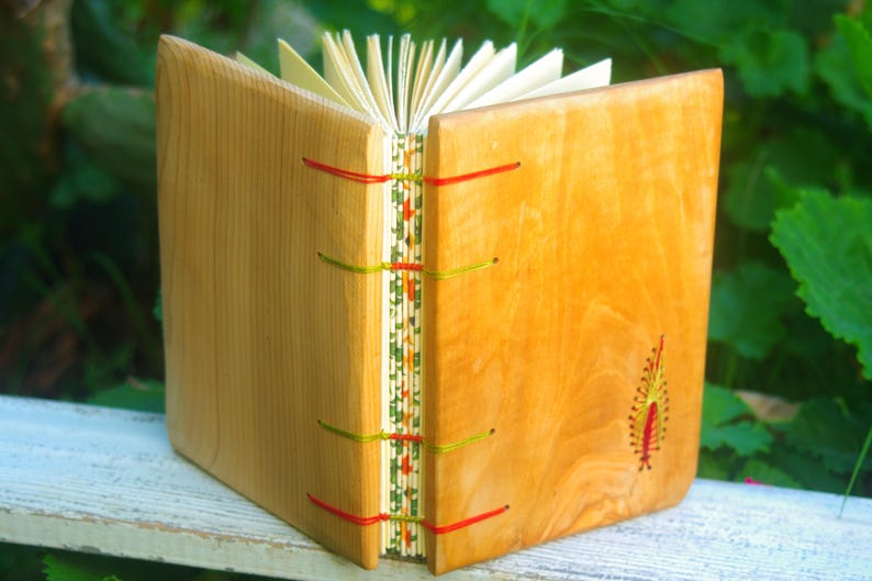 May include: A wooden journal with a natural wood cover and red, green, and yellow stitching. The journal has a decorative design on the spine and a small embroidered design on the front cover.