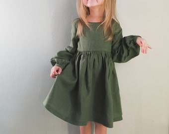 May include: A young girl wearing a green linen dress with long sleeves and a gathered skirt. She is standing in front of a light green wall and smiling at the camera.