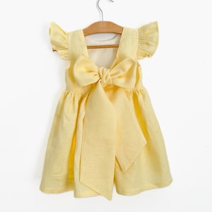 May include: A yellow linen dress with a large bow on the back. The dress has ruffled sleeves and a simple, flowy skirt.