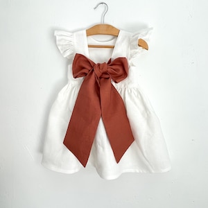 May include: A white linen dress with flutter sleeves and a large rust-colored bow. The dress is hanging on a wooden hanger against a white background. The bow is tied in the center of the dress, creating a focal point.