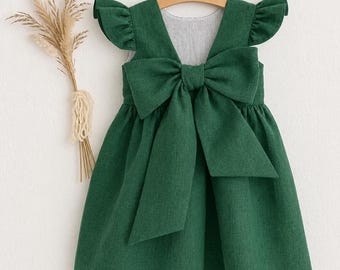 May include: A dark green sleeveless dress featuring ruffled shoulder details and a large, prominent bow centered on the back. The garment is displayed on a wooden hanger, showcasing a solid-colored fabric with a visible, textured weave.
