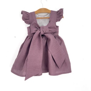 May include: A mauve linen dress with a large bow in the back. The dress has short, ruffled sleeves.