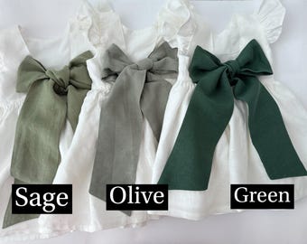 May include: Three white linen dresses with different colored sashes. The sashes are tied in a bow at the back of the dresses. The sashes are sage green, olive green, and forest green.