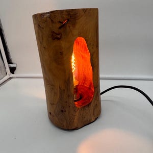May include: A wooden table lamp crafted from a section of a tree trunk. The lamp features a natural wood grain finish. An irregular opening reveals the warm glow of the light bulb inside. A black power cord extends from the base. The lamp is approximately 25 cm tall.