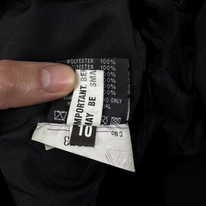 May include: A black garment interior featuring a white fabric care label with black text. The label displays 100% polyester content, safety warnings including 'IMPORTANT. BEWARE MAY BE SMALL', and a 'C No.' field. The fabric is a solid, dark black material.