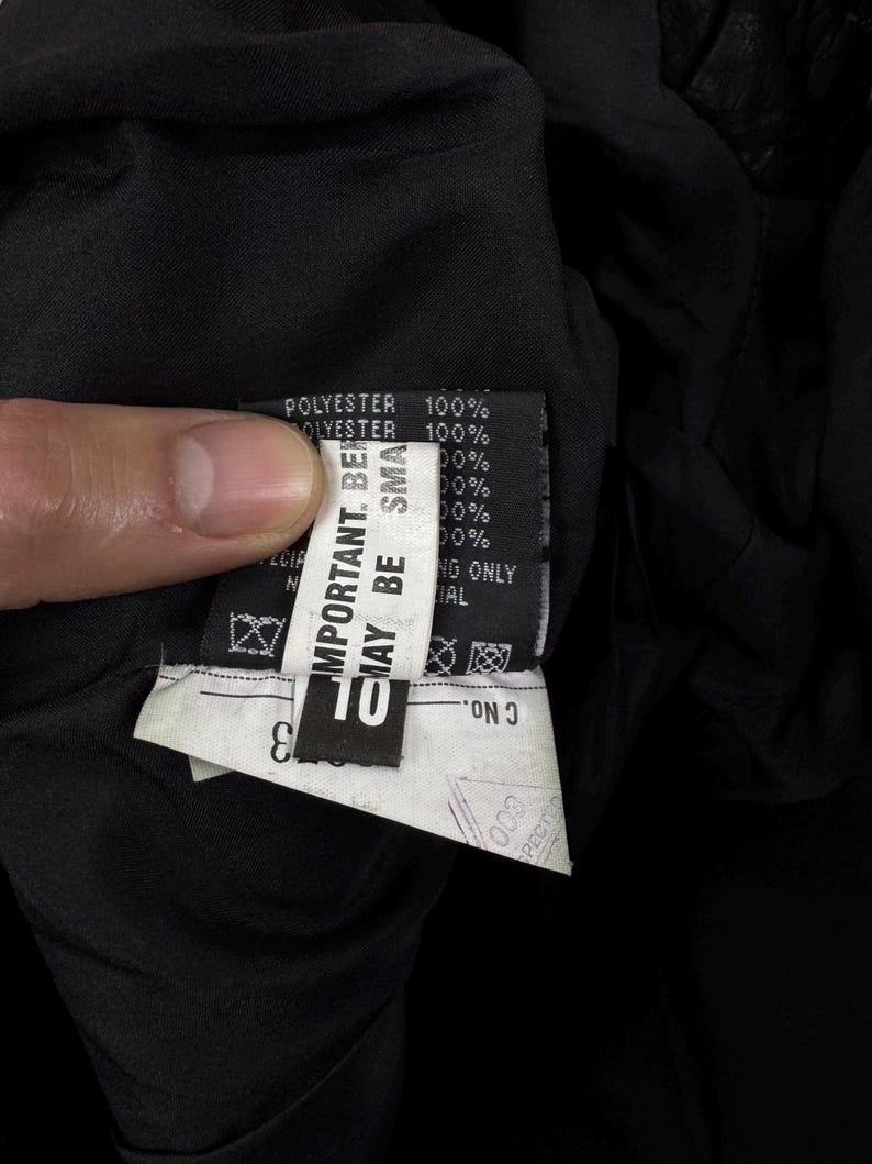 May include: A black garment interior featuring a white fabric care label with black text. The label displays 100% polyester content, safety warnings including 'IMPORTANT. BEWARE MAY BE SMALL', and a 'C No.' field. The fabric is a solid, dark black material.