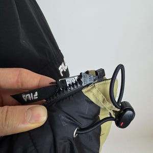 Puede incluir: Primer plano de una chaqueta negra y beige con cremallera y cord&oacute;n. La chaqueta tiene el logotipo de FILA en blanco en el tirador de la cremallera y en la manga. La cremallera es negra y tiene la marca VISLON.