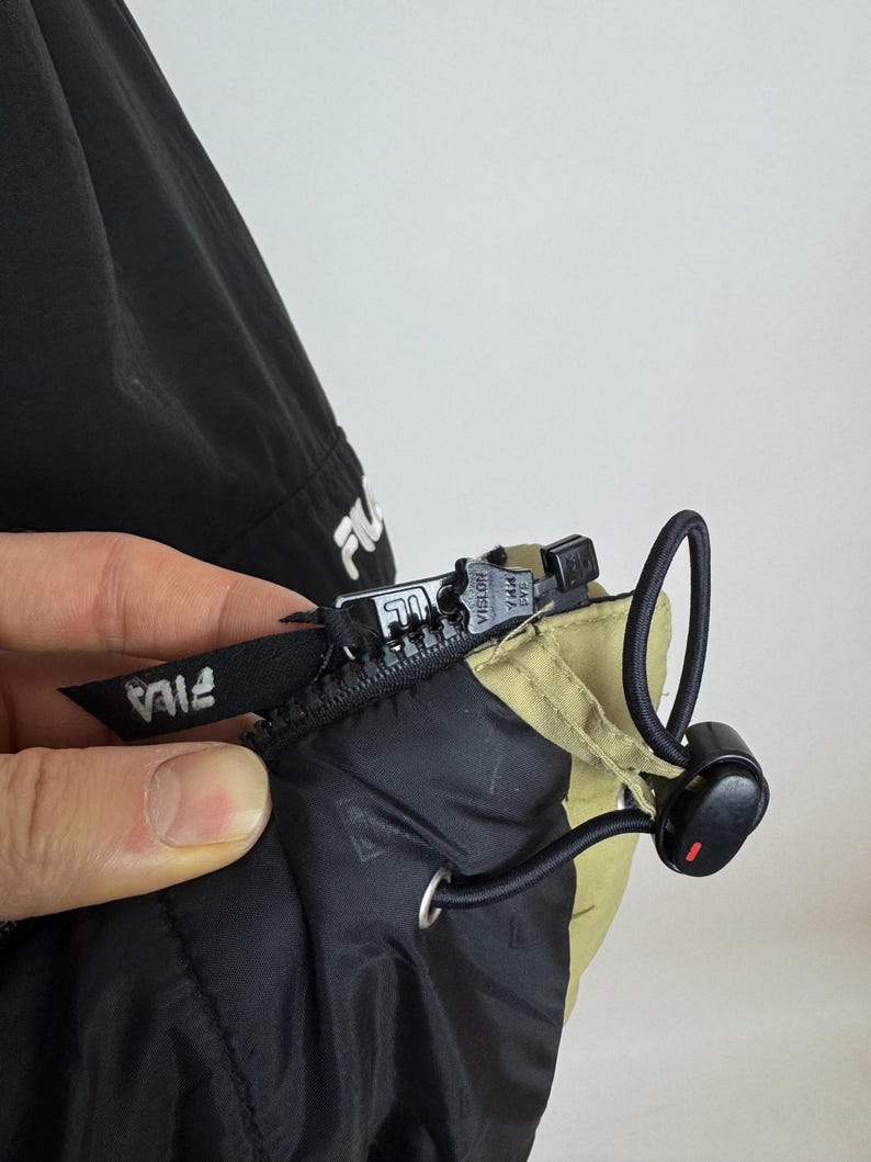 Puede incluir: Primer plano de una chaqueta negra y beige con cremallera y cord&oacute;n. La chaqueta tiene el logotipo de FILA en blanco en el tirador de la cremallera y en la manga. La cremallera es negra y tiene la marca VISLON.