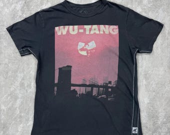 May include: Black t-shirt featuring the text "WU-TANG" in white letters above a pink and red graphic. The graphic includes a silhouette of a city skyline and the Wu-Tang Clan logo. The shirt has a crew neck and short sleeves.