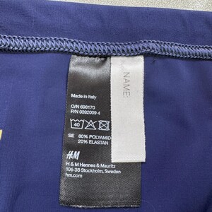 May include: Close-up of a navy blue garment with a sewn hem. A black tag with white text is attached, displaying information including "Made in Italy", fabric composition, and the H&M logo. The tag also includes care symbols.