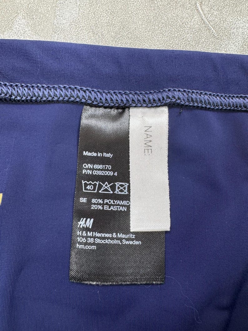 May include: Close-up of a navy blue garment with a sewn hem. A black tag with white text is attached, displaying information including "Made in Italy", fabric composition, and the H&M logo. The tag also includes care symbols.