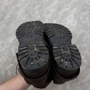 May include: A pair of brown leather work boots with thick, black rubber soles. The soles have a deep tread pattern and the CAT logo is visible. The boots show signs of wear and use.