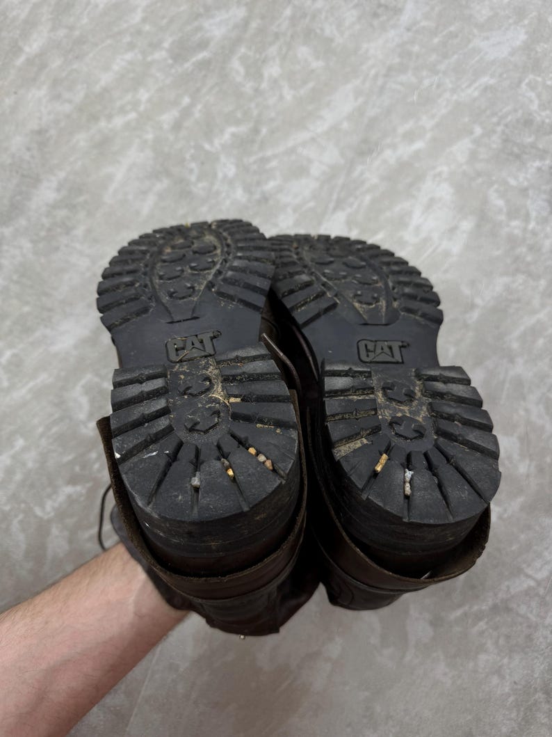 May include: A pair of brown leather work boots with thick, black rubber soles. The soles have a deep tread pattern and the CAT logo is visible. The boots show signs of wear and use.