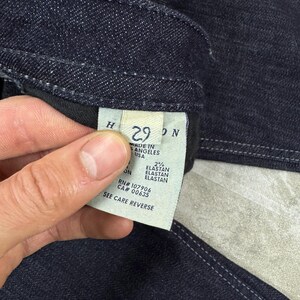 Puede incluir: Unos vaqueros de mezclilla azul oscuro con una etiqueta interior blanca. La etiqueta muestra la talla 29, el texto Made in Los Angeles USA, 2% elastan, RN# 107906, CA# 00635 y la instrucci&oacute;n See Care Reverse. La tela tiene costuras visibles.