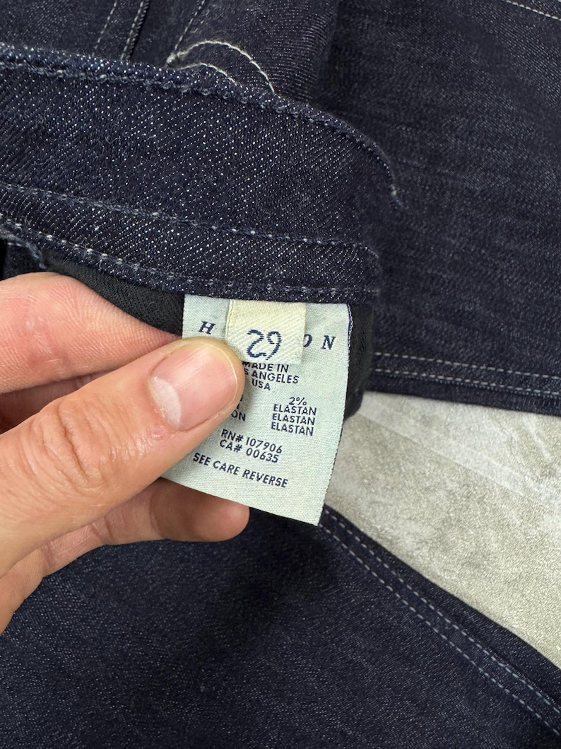Puede incluir: Unos vaqueros de mezclilla azul oscuro con una etiqueta interior blanca. La etiqueta muestra la talla 29, el texto Made in Los Angeles USA, 2% elastan, RN# 107906, CA# 00635 y la instrucci&oacute;n See Care Reverse. La tela tiene costuras visibles.