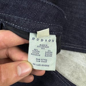 Puede incluir: Una prenda de mezclilla azul oscuro con una etiqueta de cuidado blanca en el interior. La etiqueta dice HUDSON, MADE IN LOS ANGELES USA, 98% COTTON, 2% ELASTAN, RN# 107906, CA# 00635 y SEE CARE REVERSE. La mezclilla tiene costuras blancas visibles.