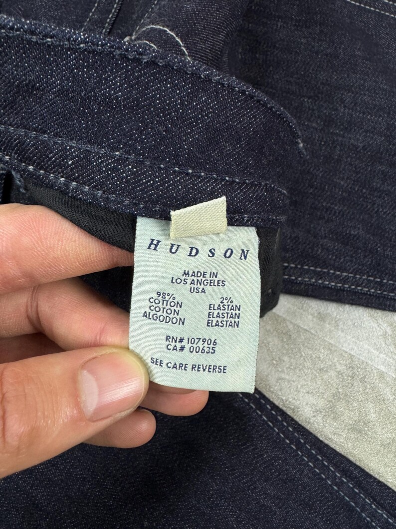 Puede incluir: Una prenda de mezclilla azul oscuro con una etiqueta de cuidado blanca en el interior. La etiqueta dice HUDSON, MADE IN LOS ANGELES USA, 98% COTTON, 2% ELASTAN, RN# 107906, CA# 00635 y SEE CARE REVERSE. La mezclilla tiene costuras blancas visibles.